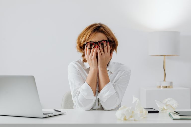 Woman with hands on face, sitting at desk with laptop and tissues, feeling stressed, mental load.