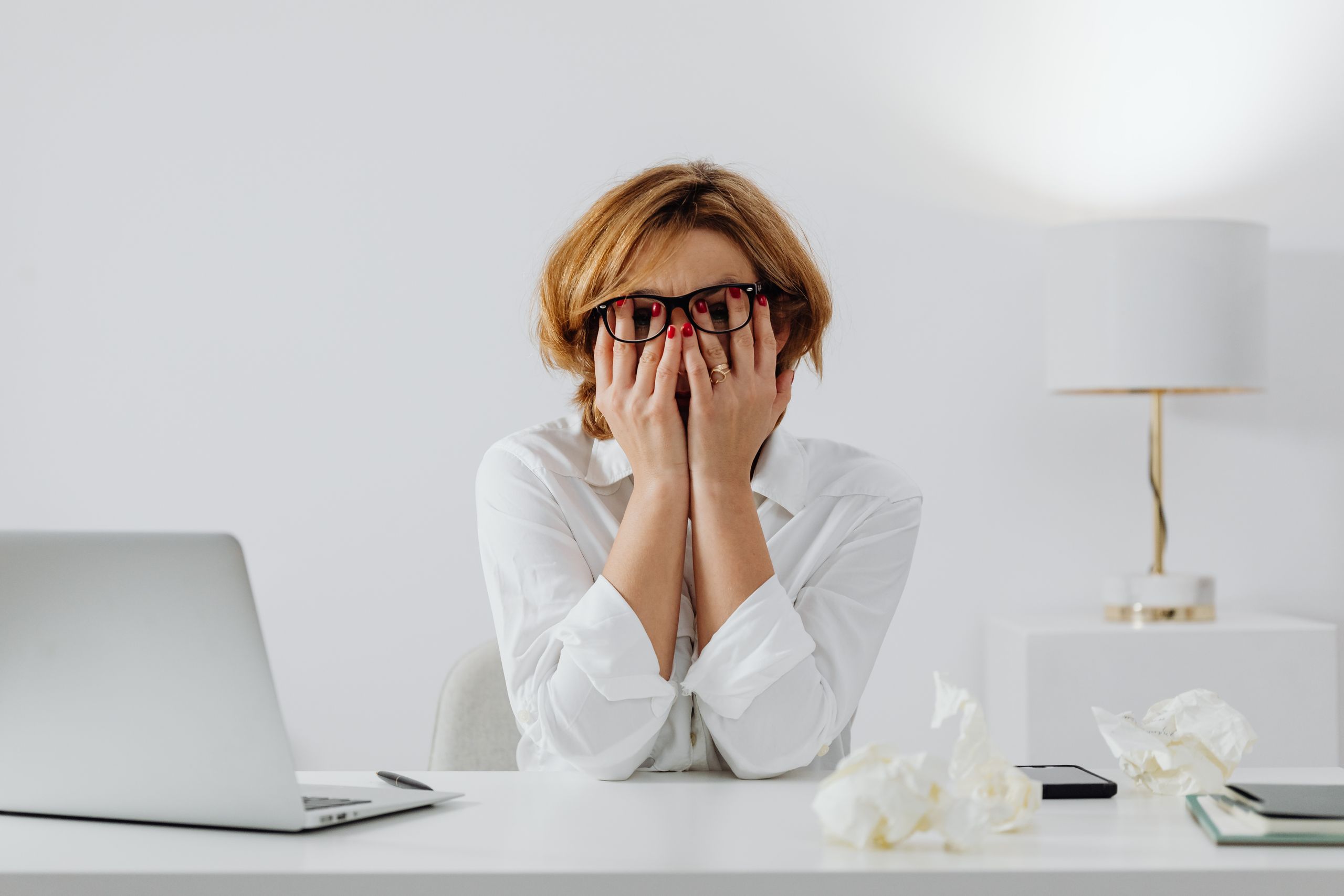 Woman with hands on face, sitting at desk with laptop and tissues, feeling stressed, mental load.
