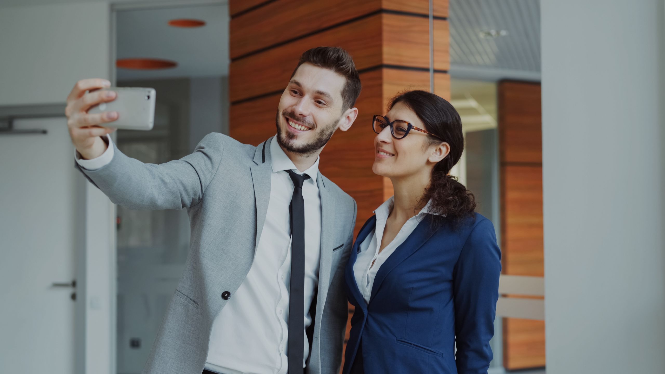 Two business colleagues smiling while taking a selfie indoors at the office.