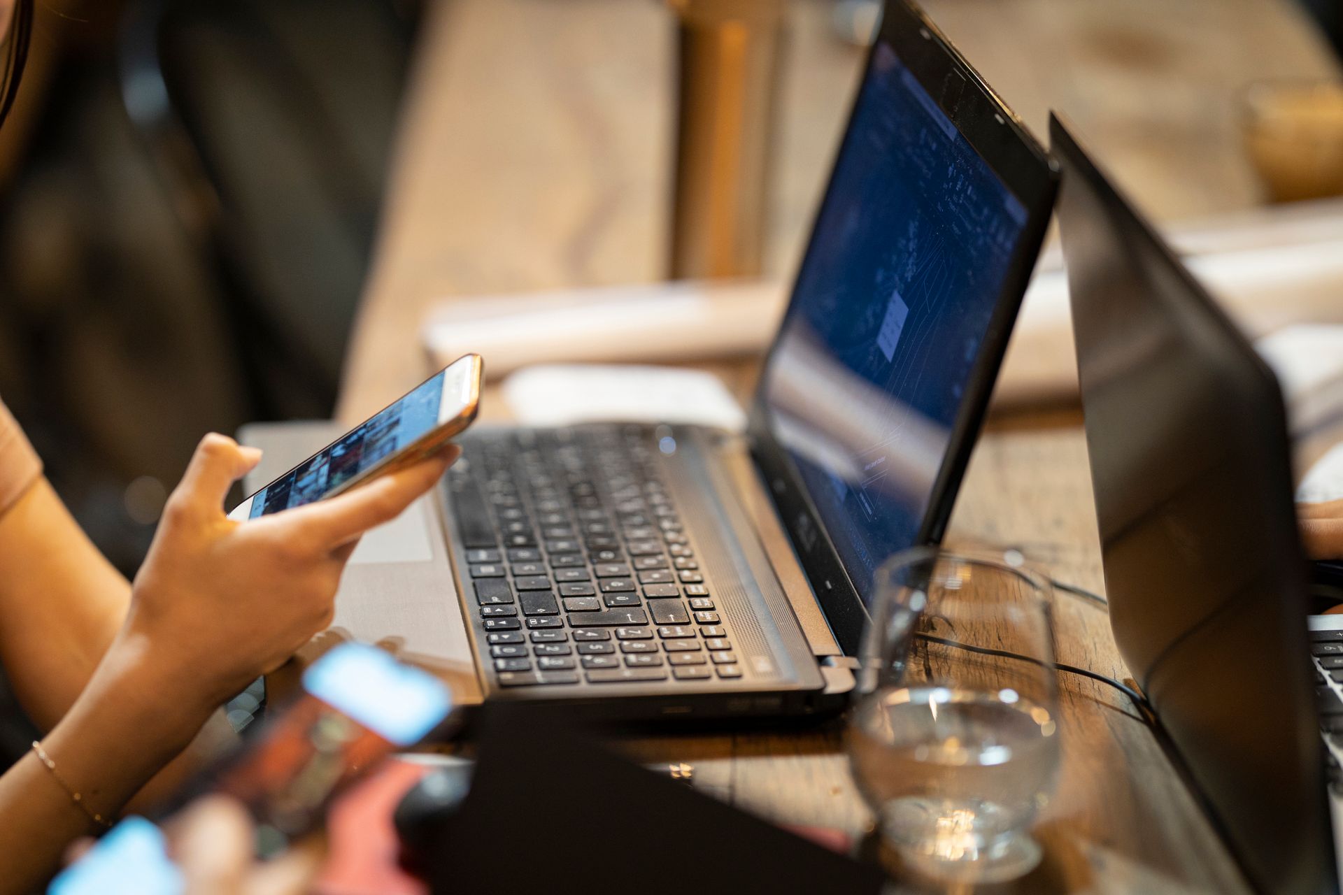 A person holding a smartphone beside an open laptop on a wooden table