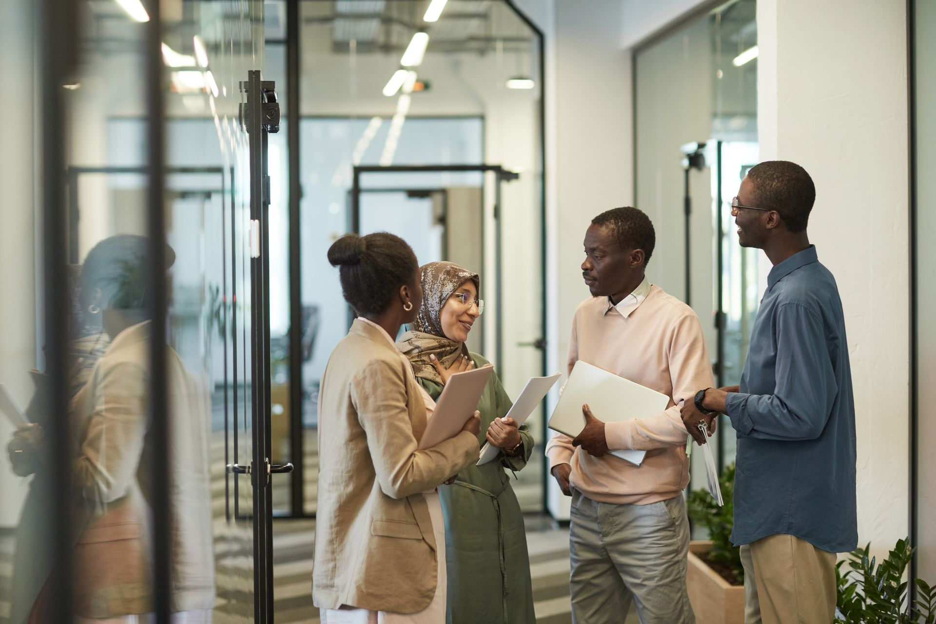 Four colleagues holding folders and a laptop talking together in a glass-walled office corridor