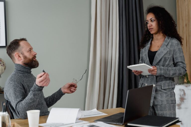A manager sitting at a desk talking with a colleague who stands holding a tablet in an office