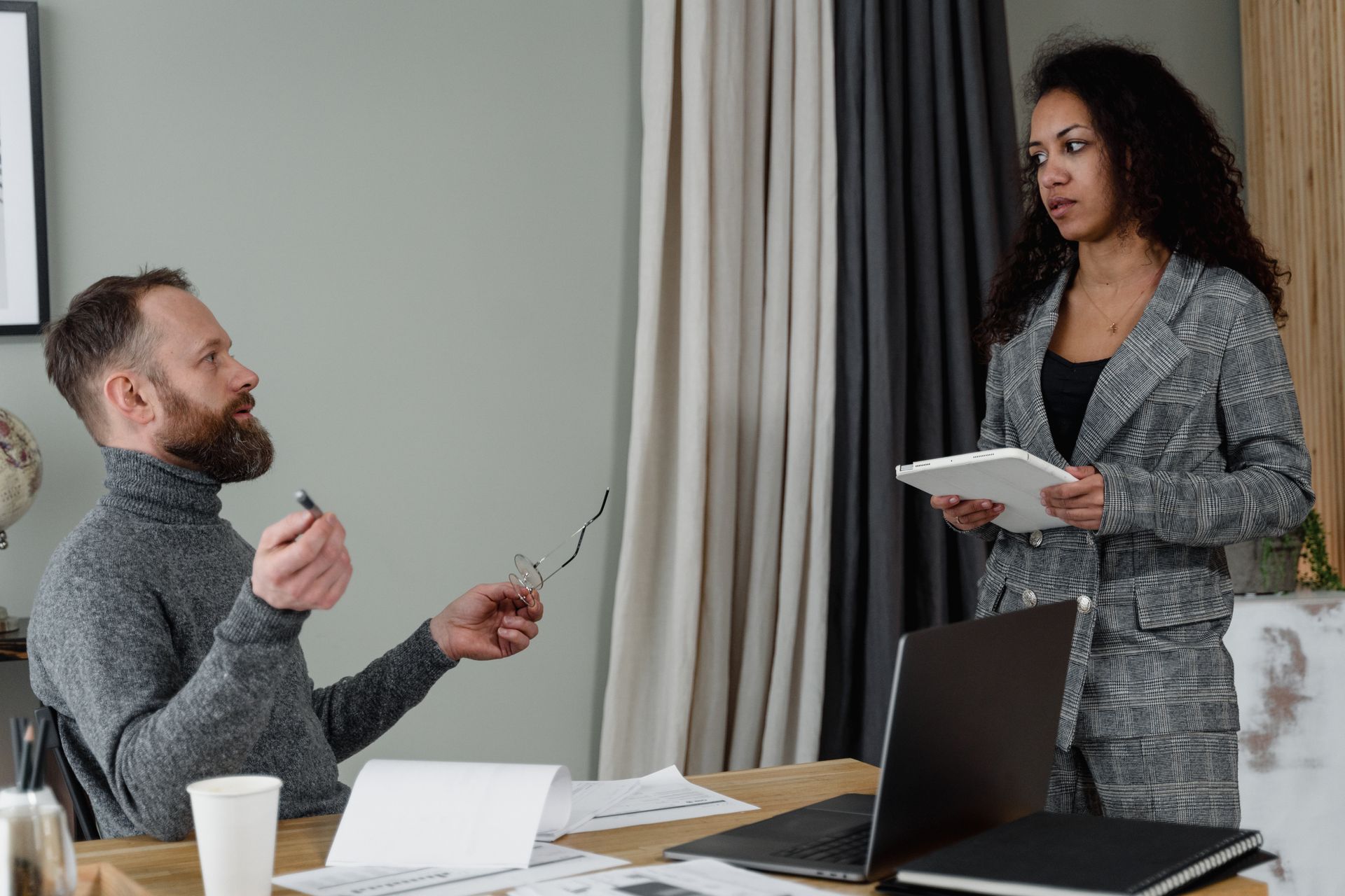 A manager sitting at a desk talking with a colleague who stands holding a tablet in an office