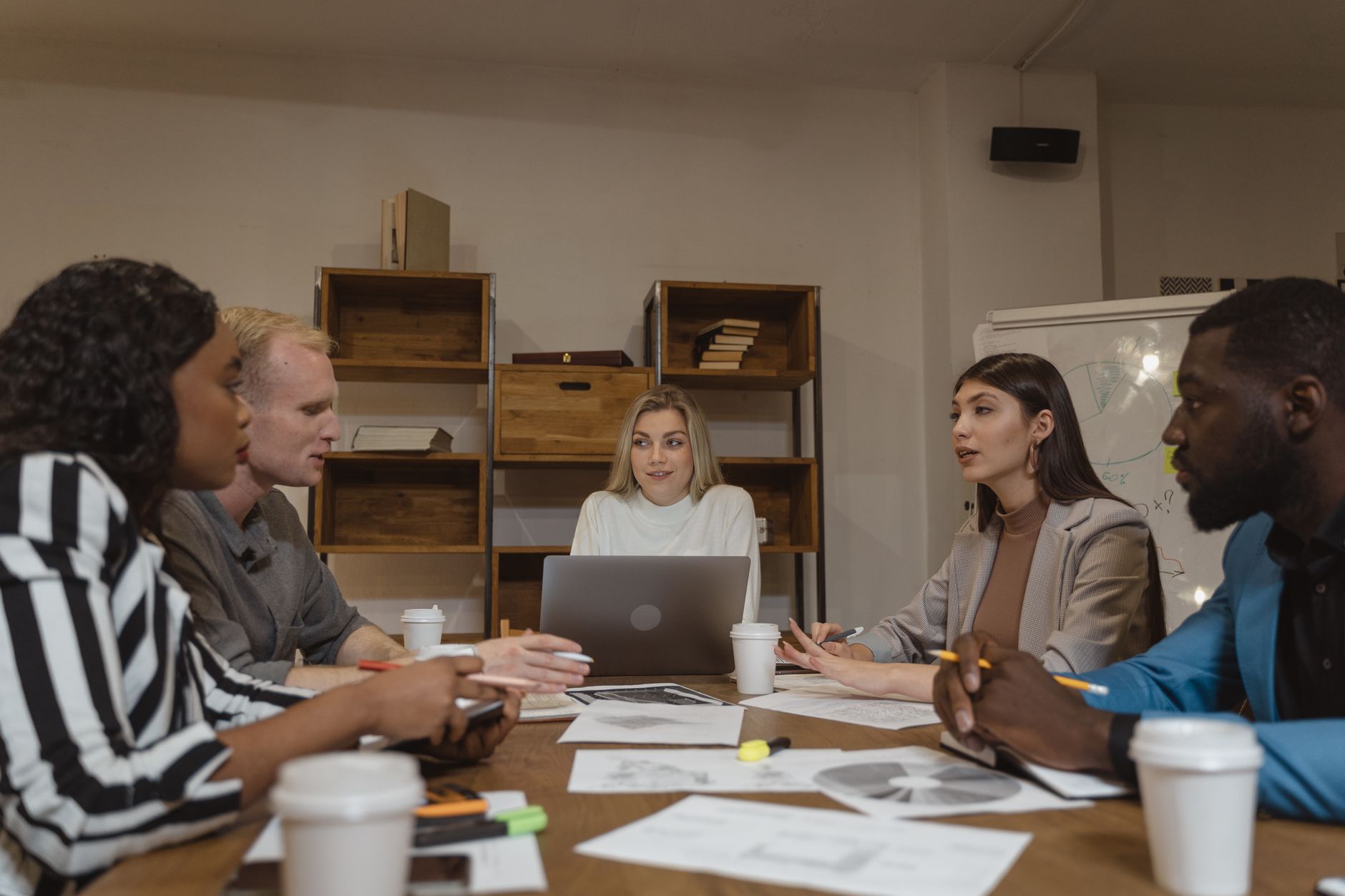 Colleagues talking around a meeting table with laptops, notes and coffee cups in an office